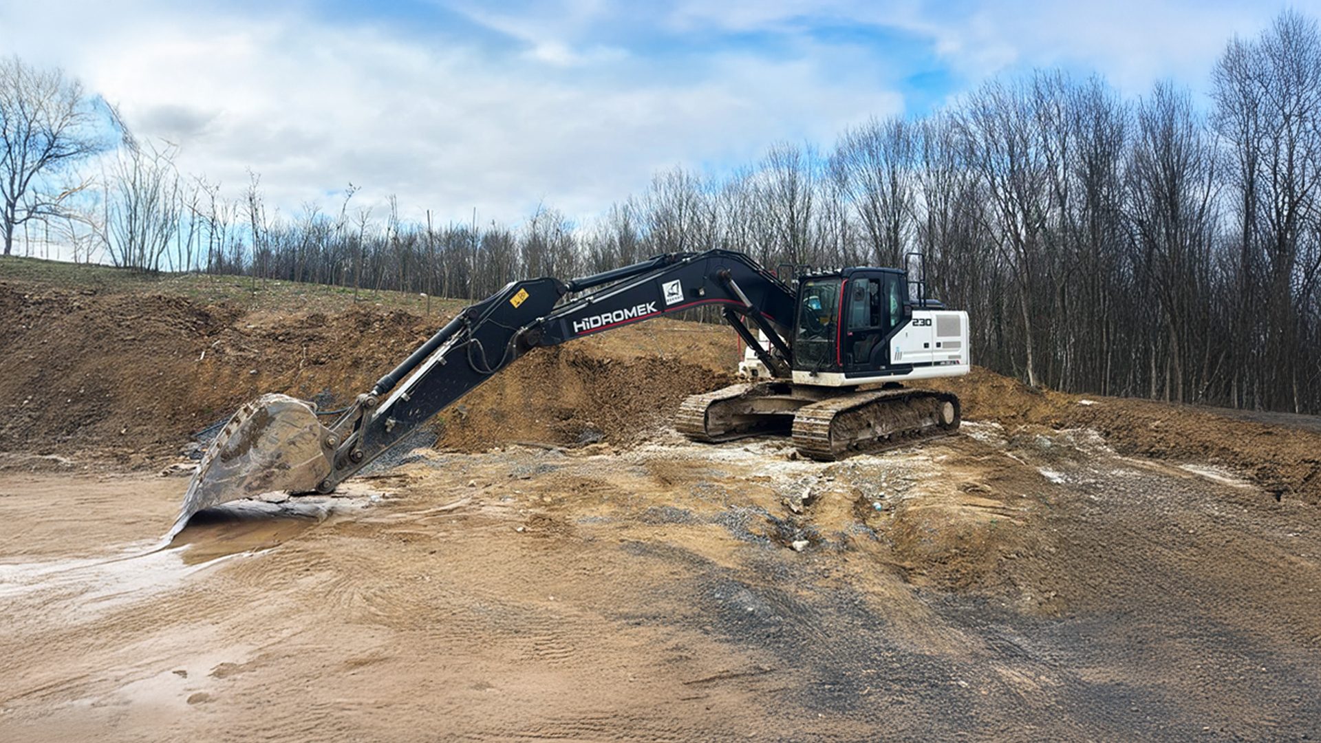 Black and white HİDROMEK excavator on a dirt construction site under a cloudy sky with bare trees.