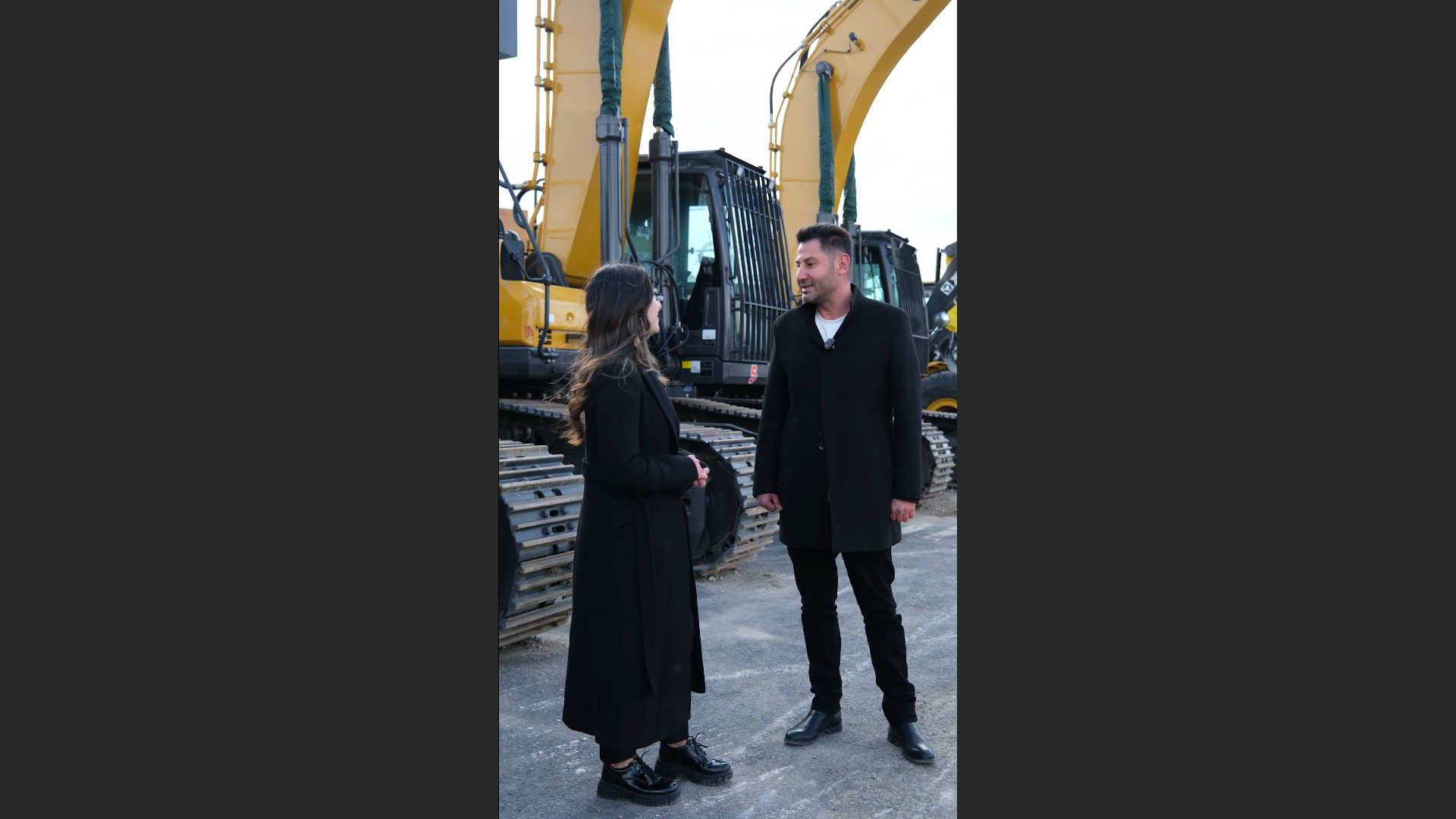 Two people talking in black coats, standing in front of large yellow excavators outdoors.