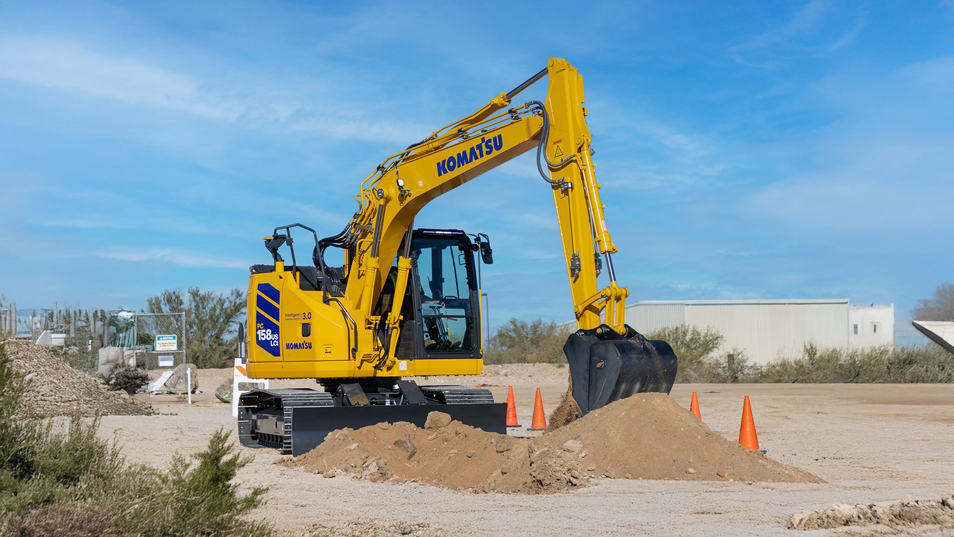 A yellow Komatsu excavator on a dirt construction site with piles of dirt and orange cones under a blue sky.