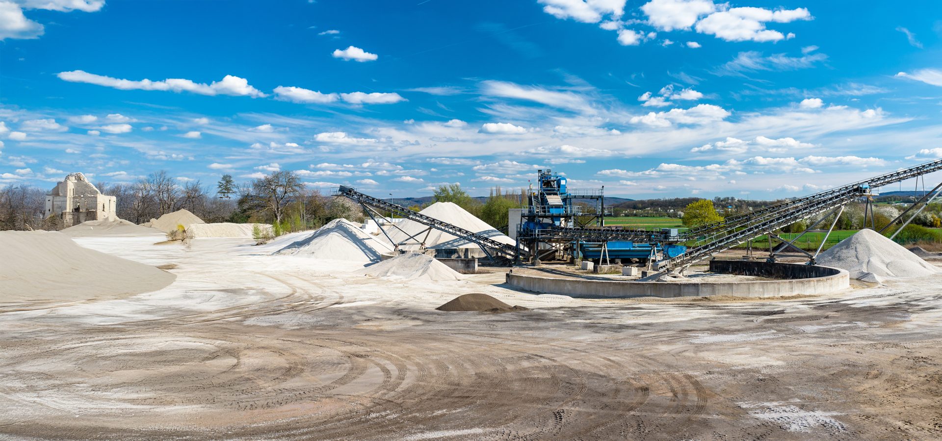 Sand and gravel quarry featuring blue processing machinery, conveyor belts, material piles, and blue sky.