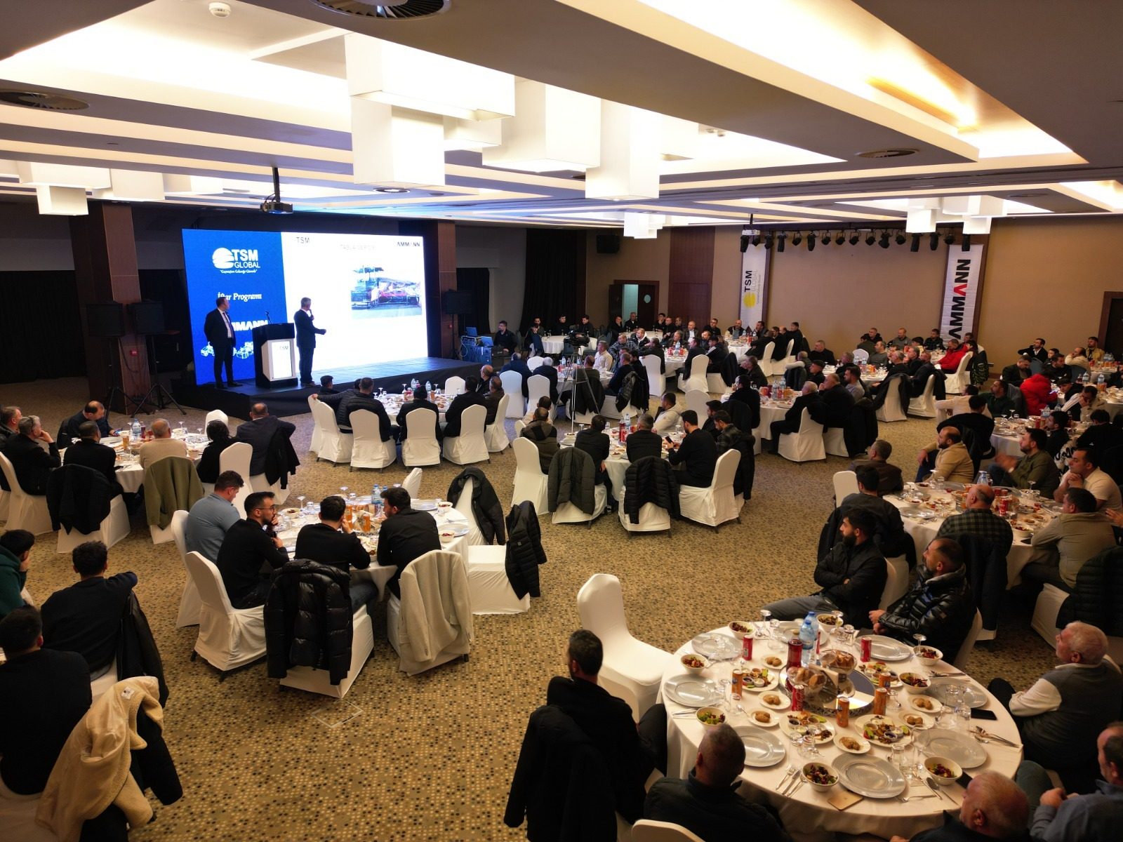 An indoor event with speakers presenting on a stage with a large screen to an audience seated at tables.