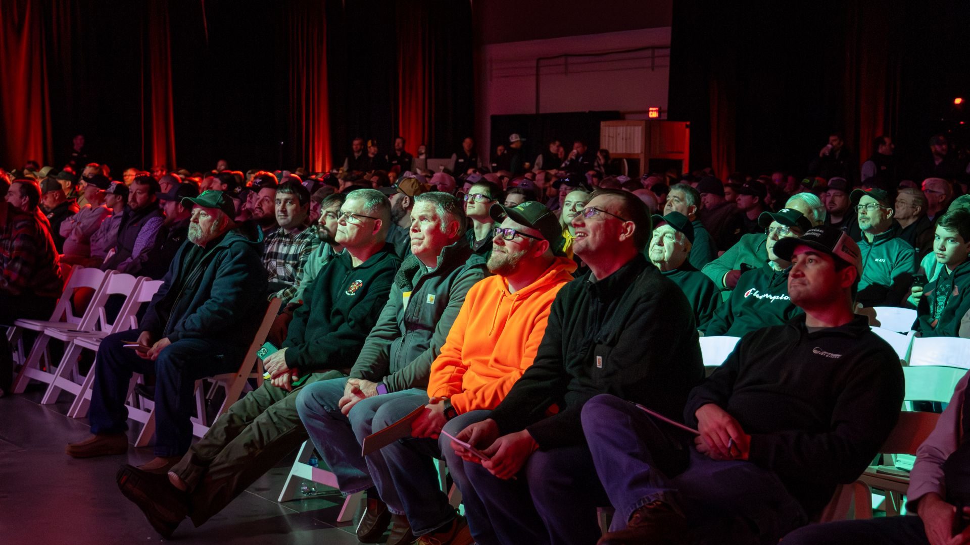 Audience of men watching an event intently in a dimly lit room with red and green lights.