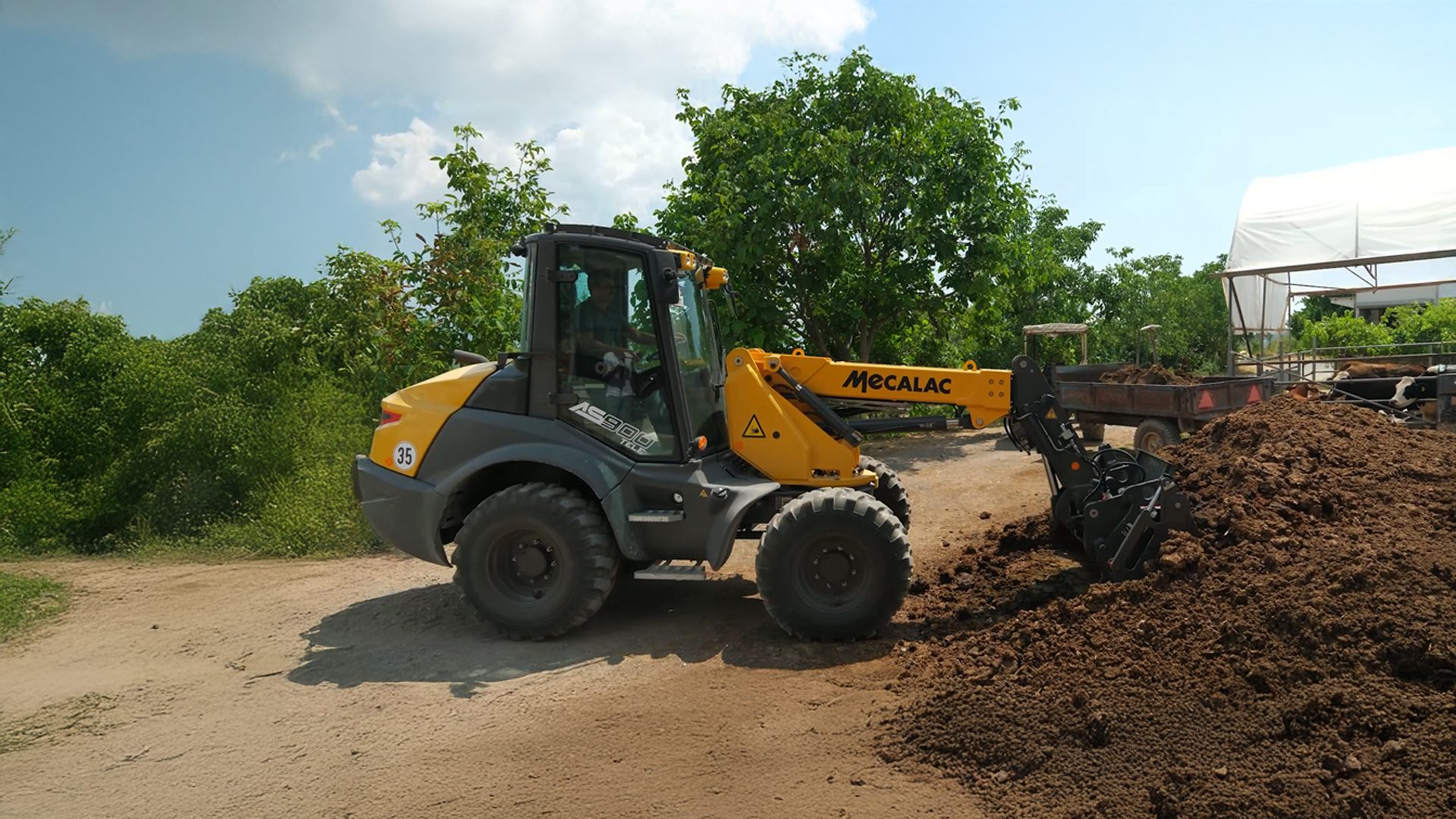 Mecalac AS900 loader moving manure on a farm with cows and trees.