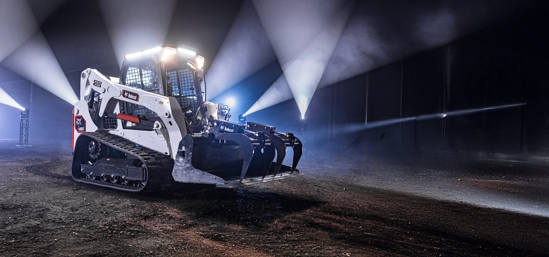White Bobcat T650 compact track loader with a grapple attachment, illuminated by bright spotlights.