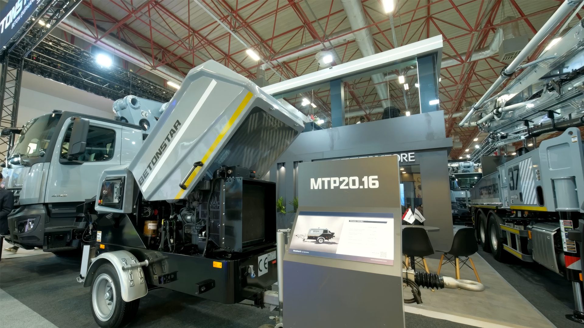 Grey concrete pump truck with open hood and display stand at an exhibition.