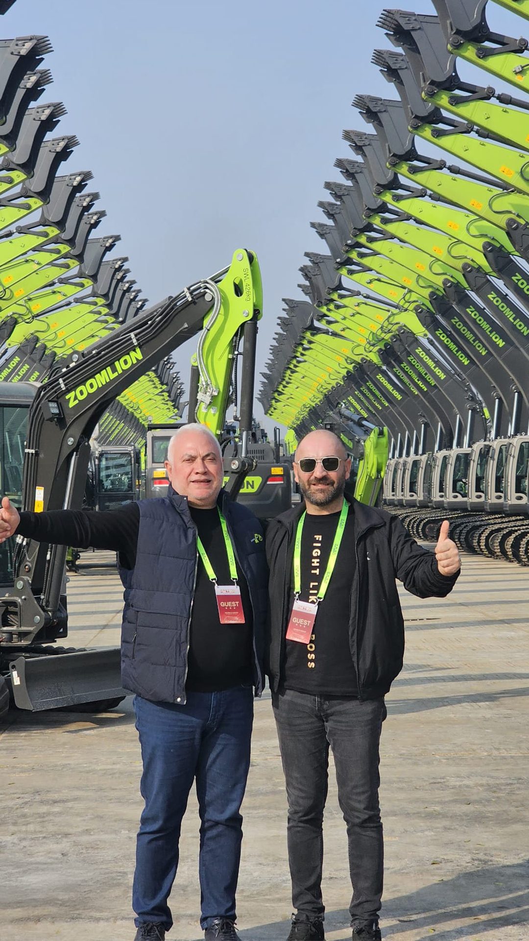 Two men give thumbs-up in front of rows of green ZOOMLION excavators.