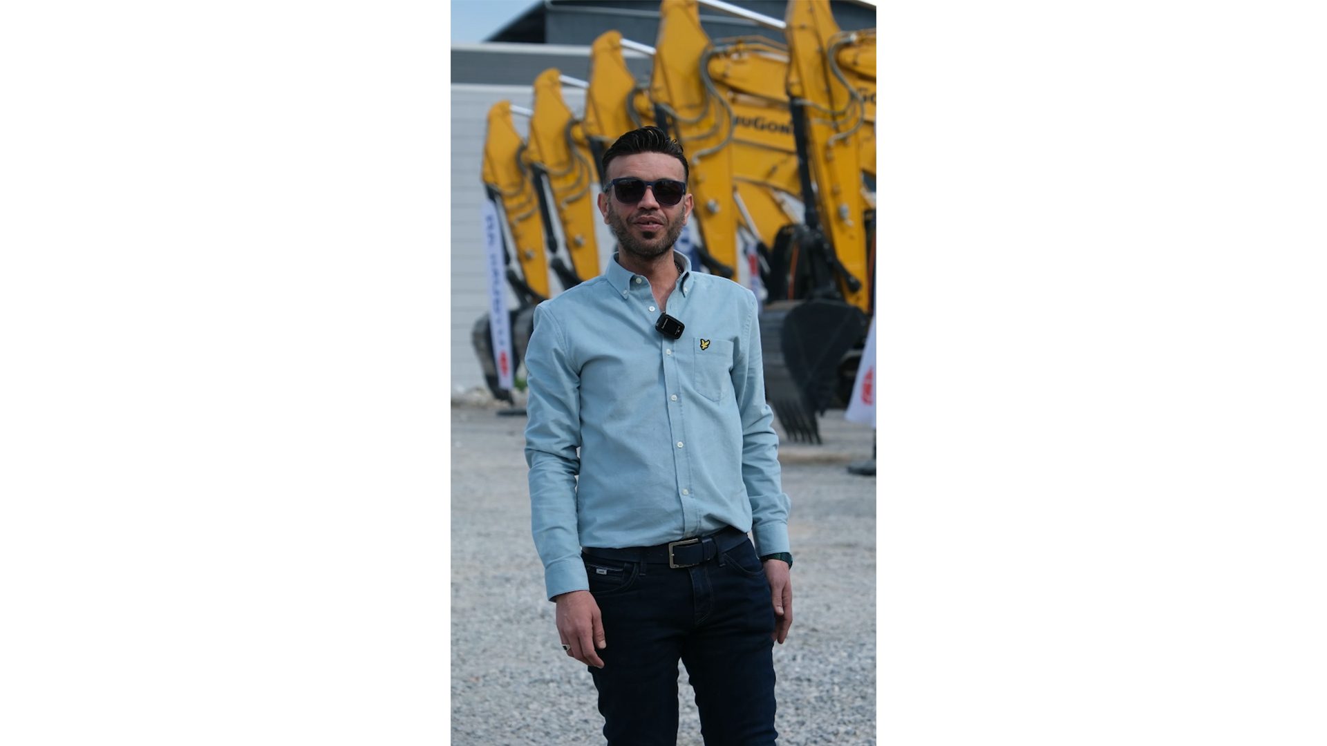 A man in sunglasses and a light blue shirt stands casually outdoors, with yellow excavators in the background.