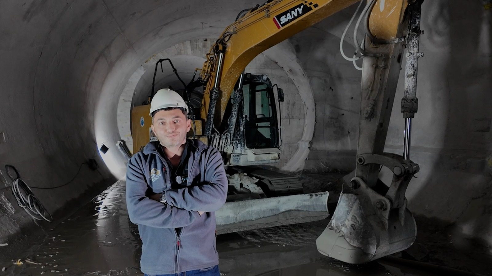 Man in a hard hat, arms crossed, stands in a concrete tunnel with a large excavator behind him.