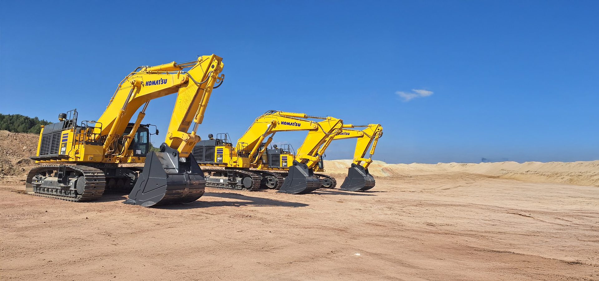 Three yellow Komatsu excavators on a sandy construction site under a clear blue sky.