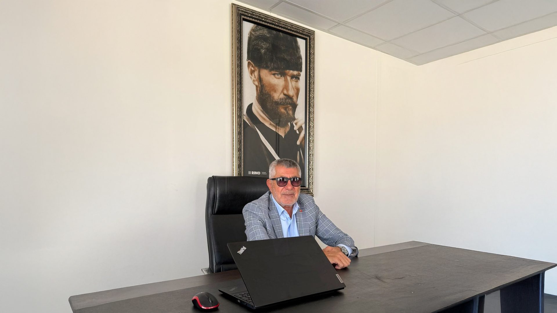 Businessman in sunglasses at desk with laptop, Atatürk portrait behind.