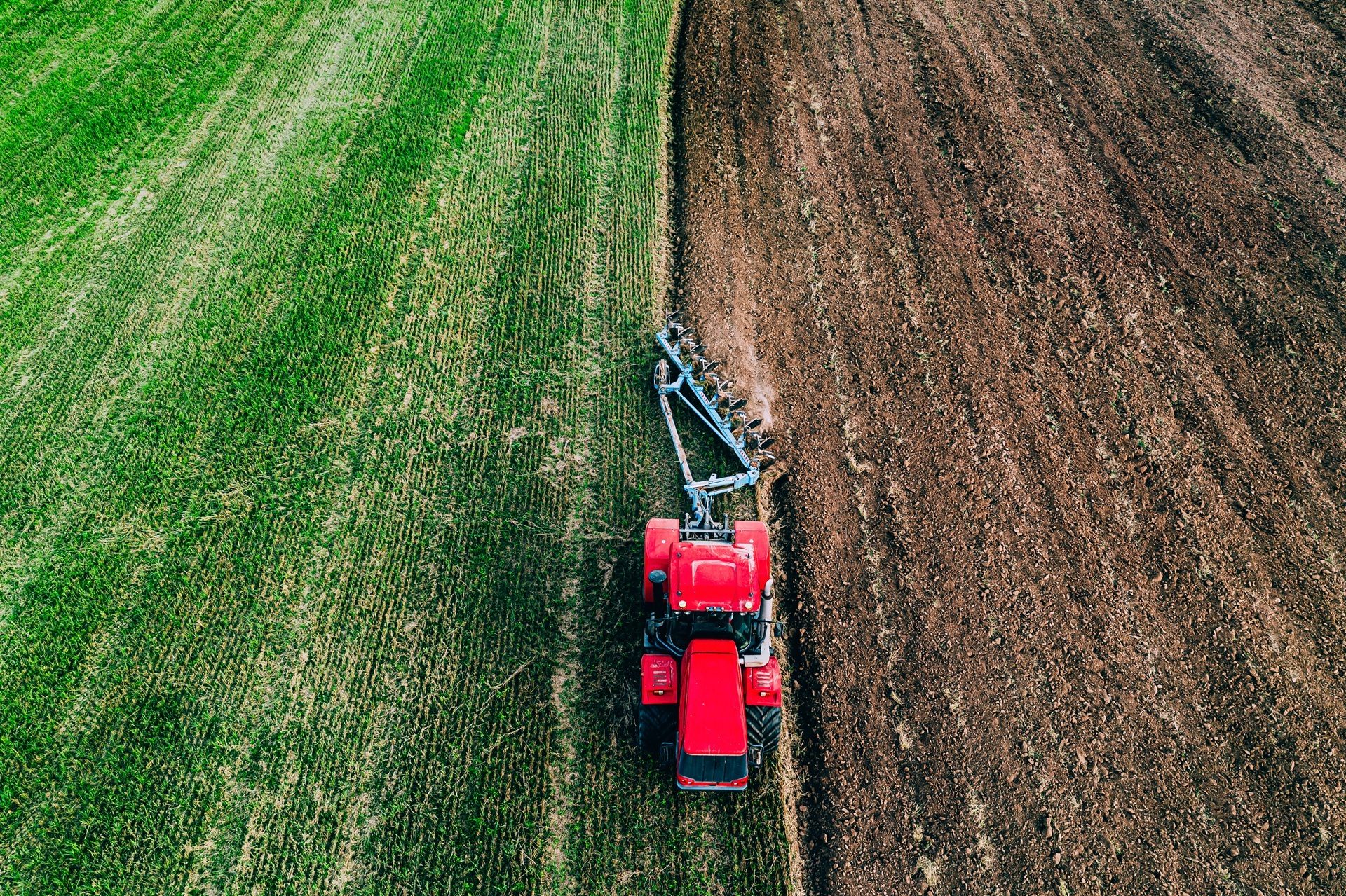 Aerial view of a red tractor plowing a field, dividing it into green and tilled brown earth.