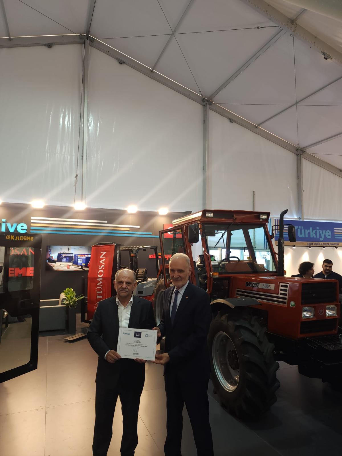 Two men in suits holding a certificate in front of a red TÜMOSAN tractor at an exhibition.