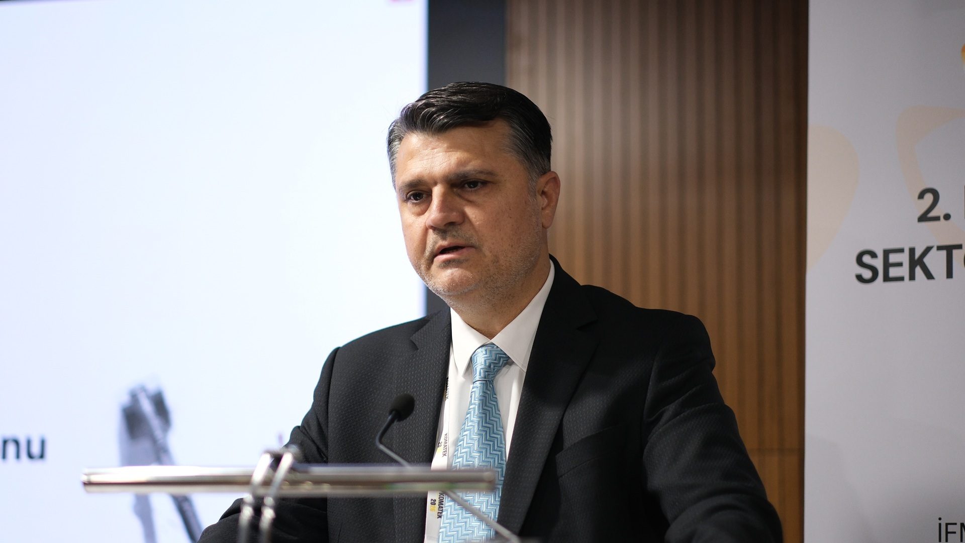 A man in a dark suit and light blue tie speaks at a podium.