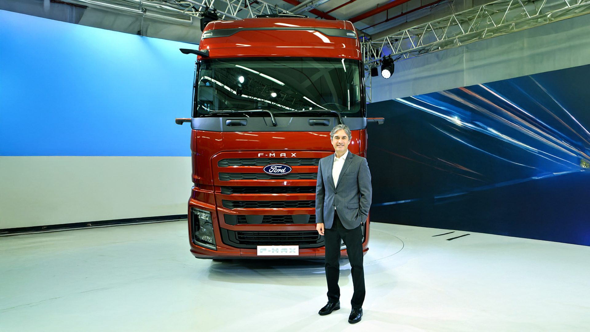 Man in a suit standing proudly in front of a new red Ford F-Max truck at its debut event.
