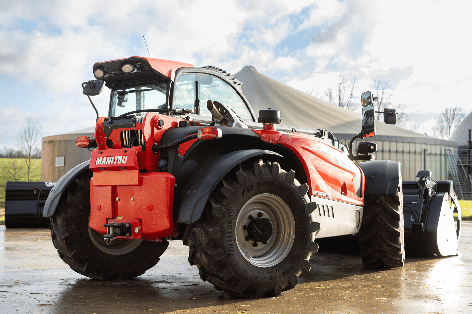 Red Manitou telehandler with large tires and shovel attachment parked outdoors near industrial silos.