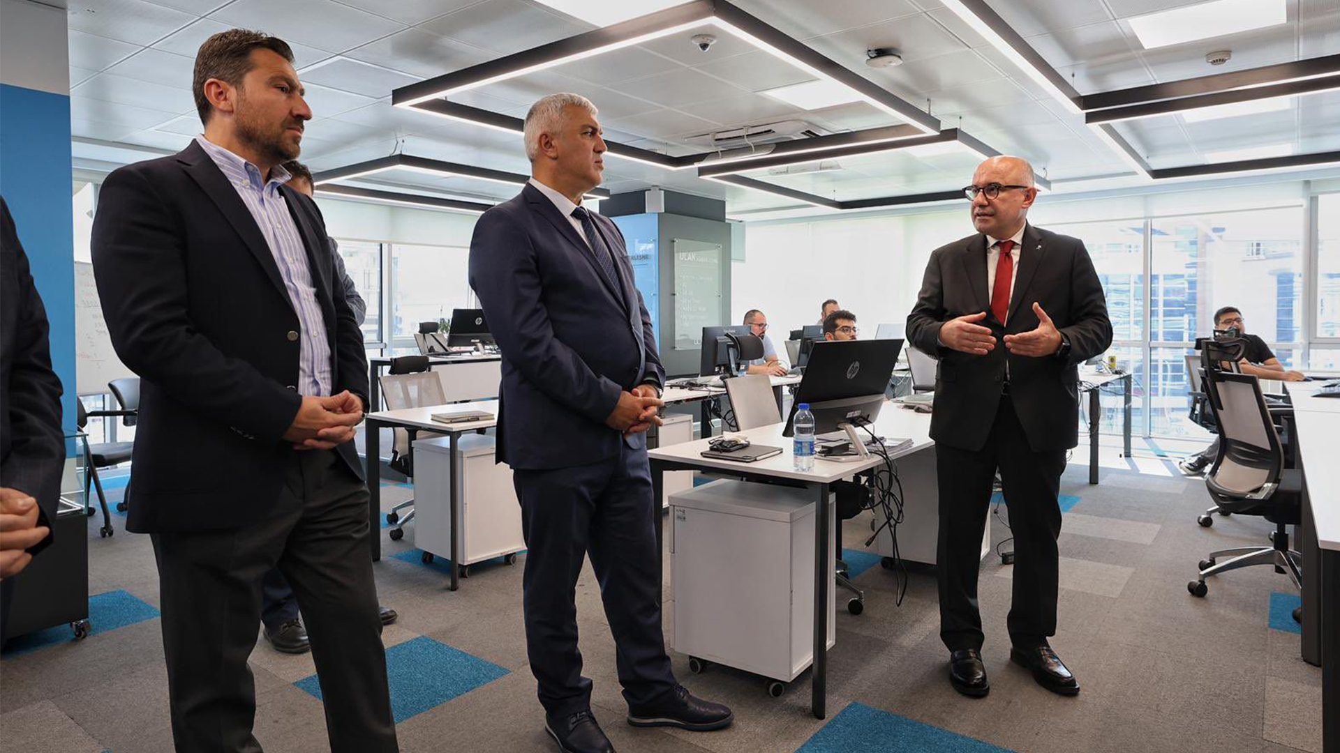 Businessmen in a modern office, one speaking to others who are listening.