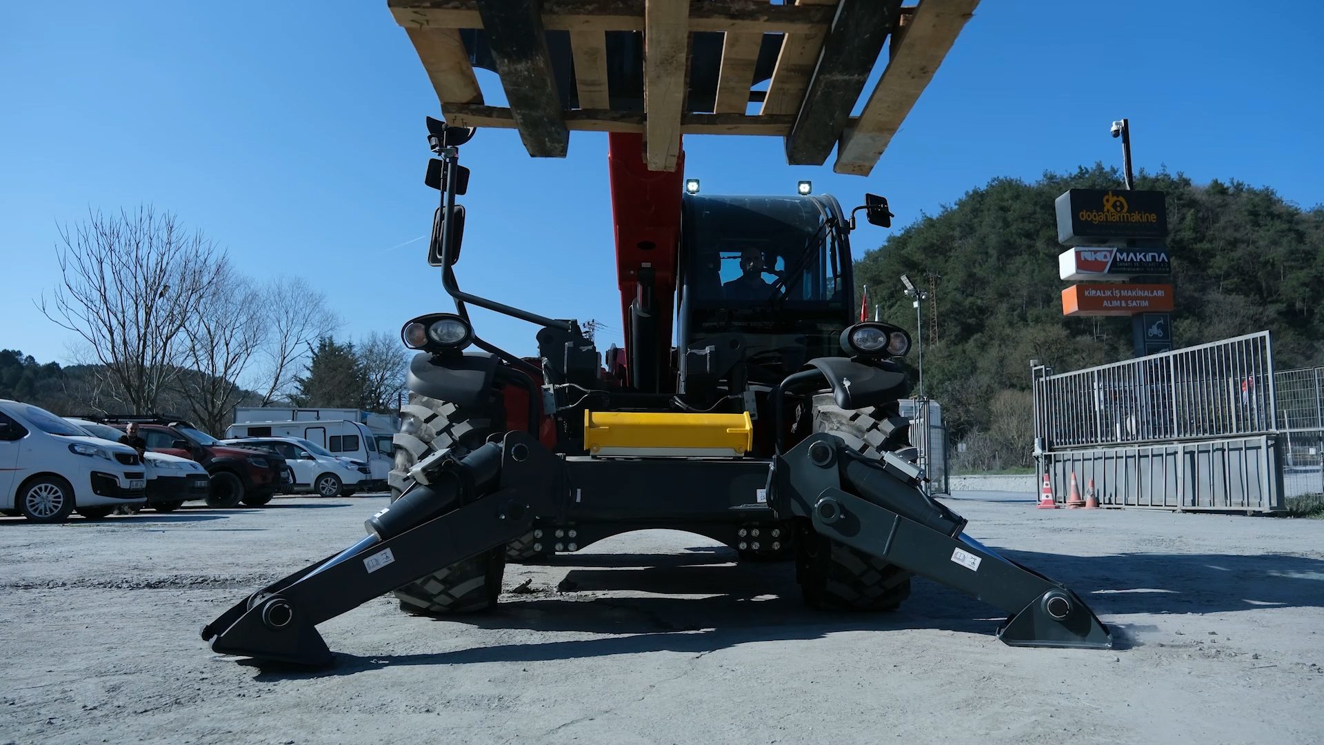 A telehandler with extended outriggers, holding wooden pallets, parked in a lot with cars.