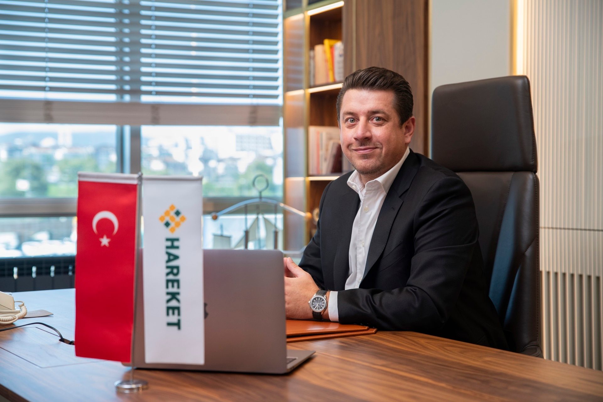 Smiling man in a suit sits at an office desk with a Turkish flag and a "HAREKET" flag.