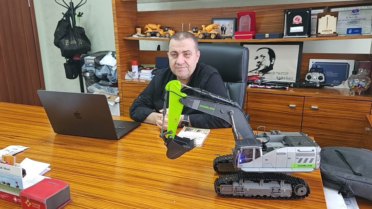 A man sits at a wooden office desk with a laptop and a large Zoomlion toy excavator model.