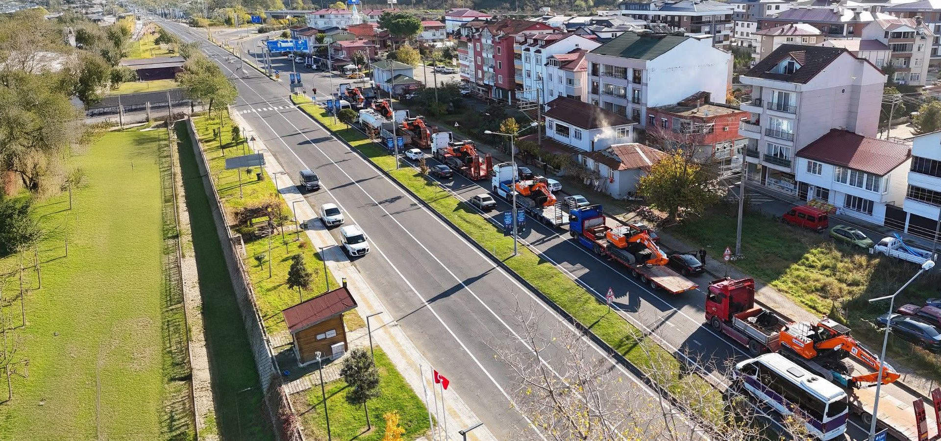 Long line of trucks carrying excavators parked on a roadside in a town with buildings.