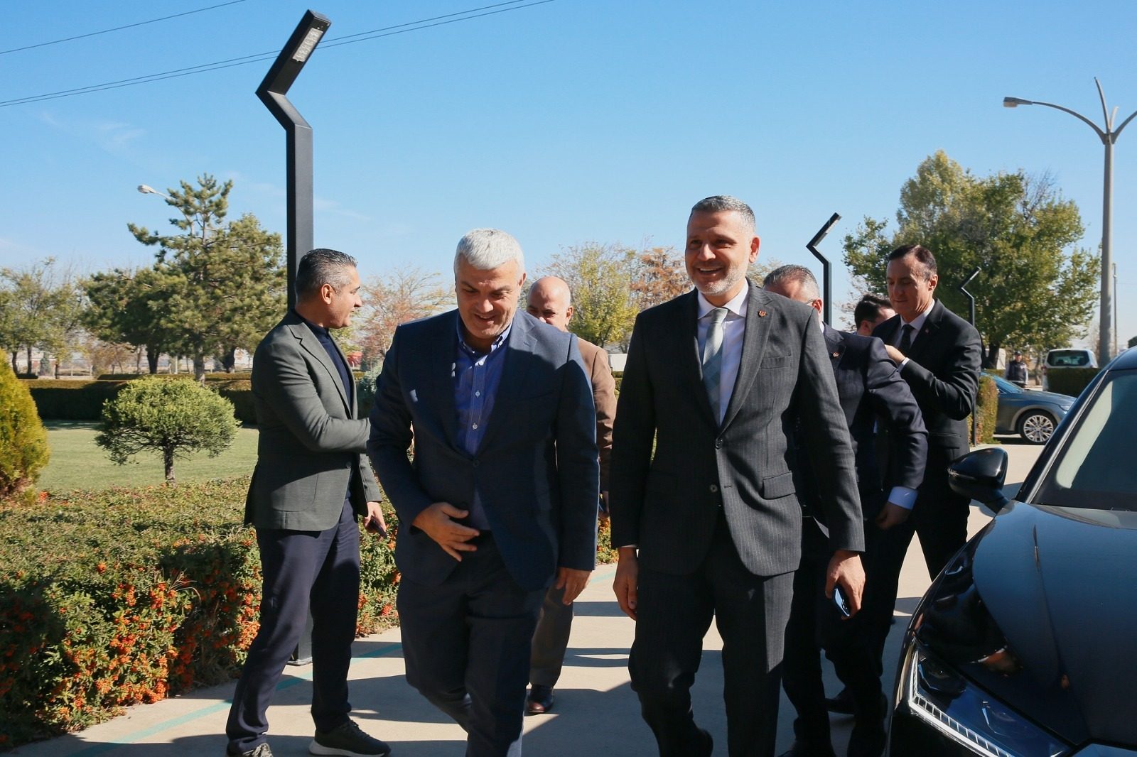 Two smiling men in suits walk with a group outdoors on a sunny day, with greenery and a car.