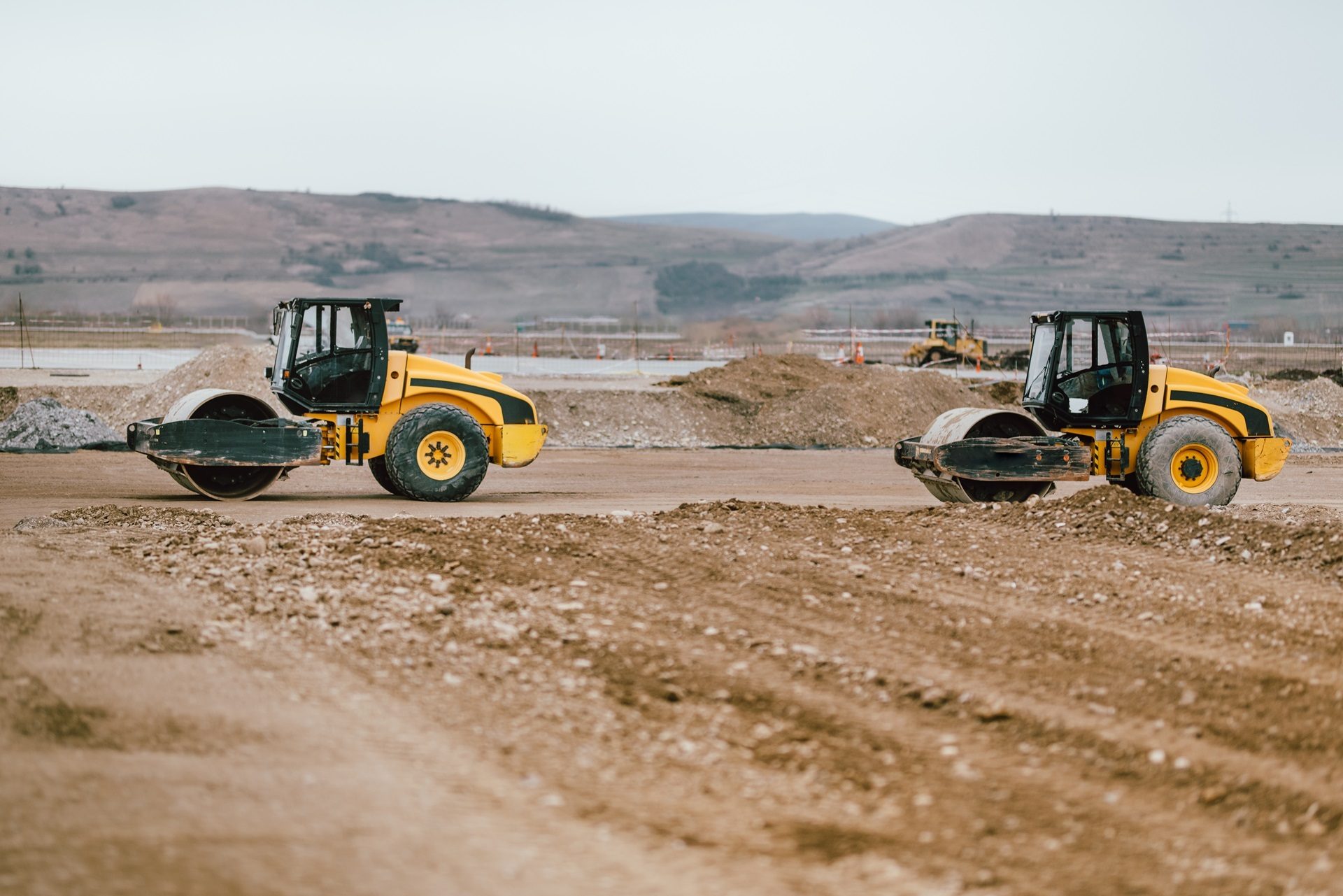 Two yellow road rollers on a construction site with dirt and distant hills.