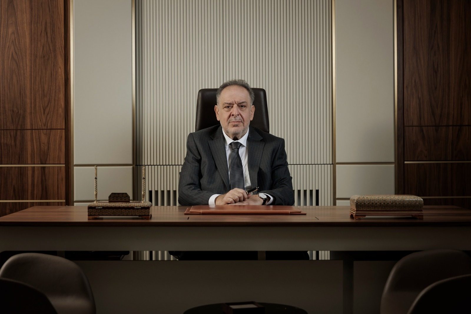 Man in a dark suit sitting at a large wooden desk in a modern office.