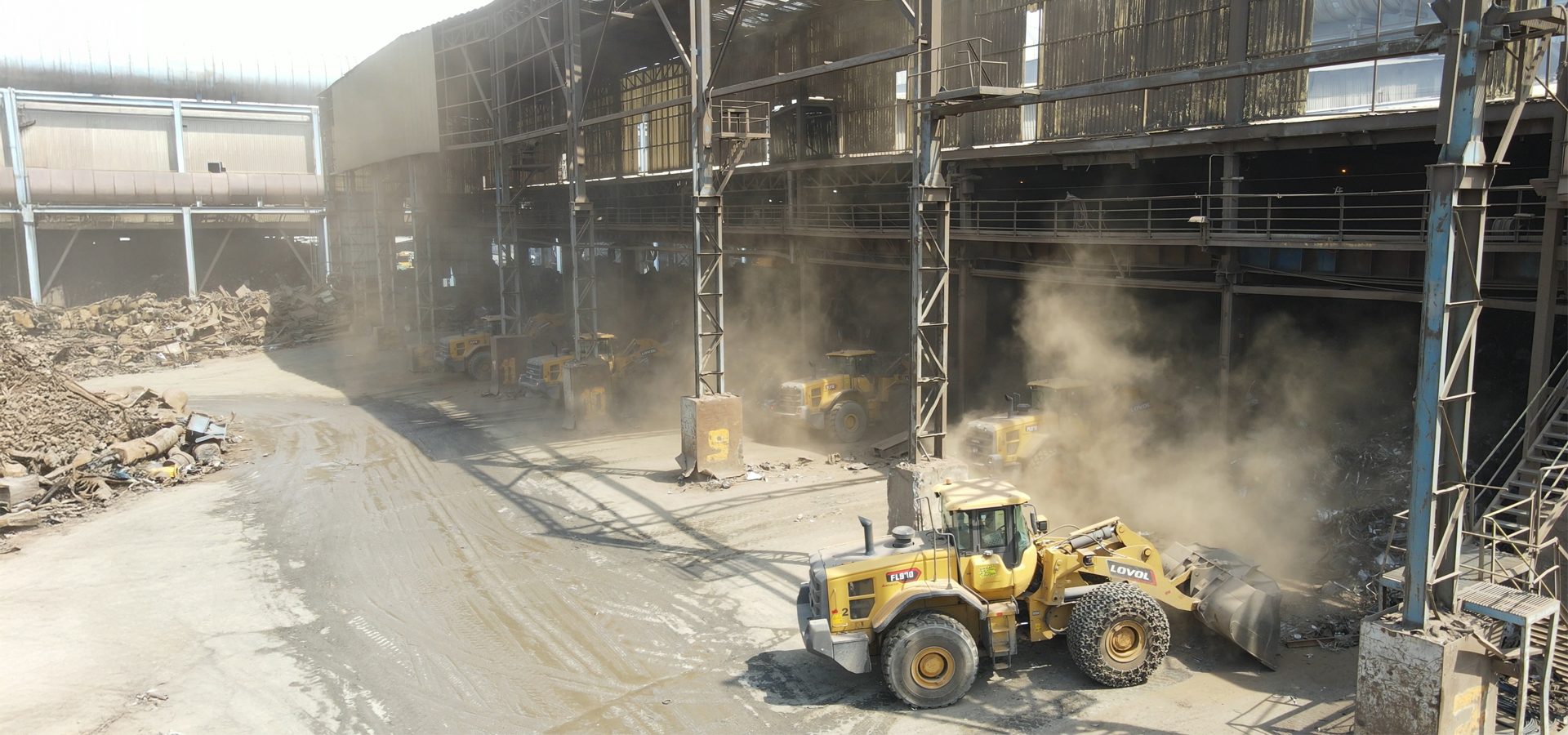 Dusty industrial yard with yellow loaders moving scrap metal.