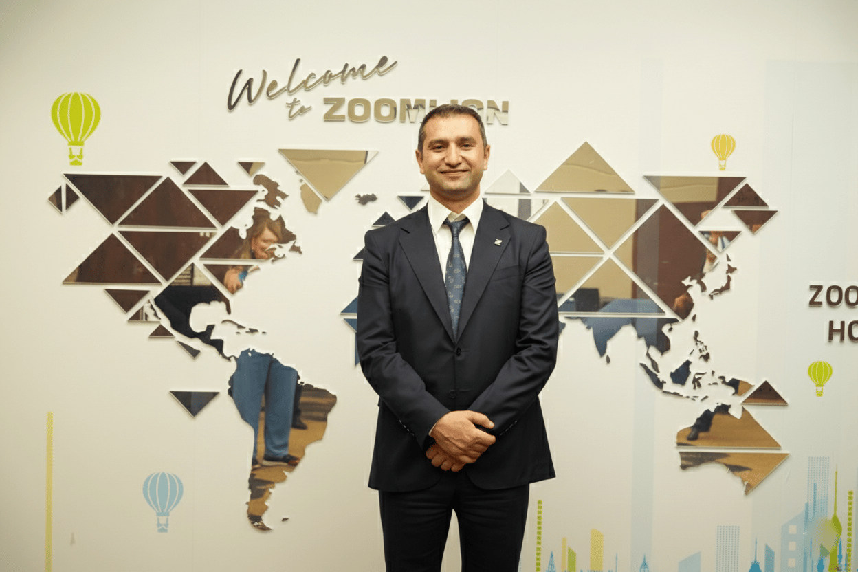 Man in suit smiles in front of 'Welcome to ZOOMLION' sign and mirrored world map.