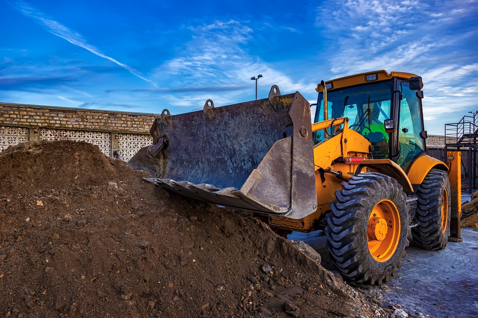 A yellow backhoe with its large metal bucket positioned over a pile of brown dirt under a bright blue sky.
