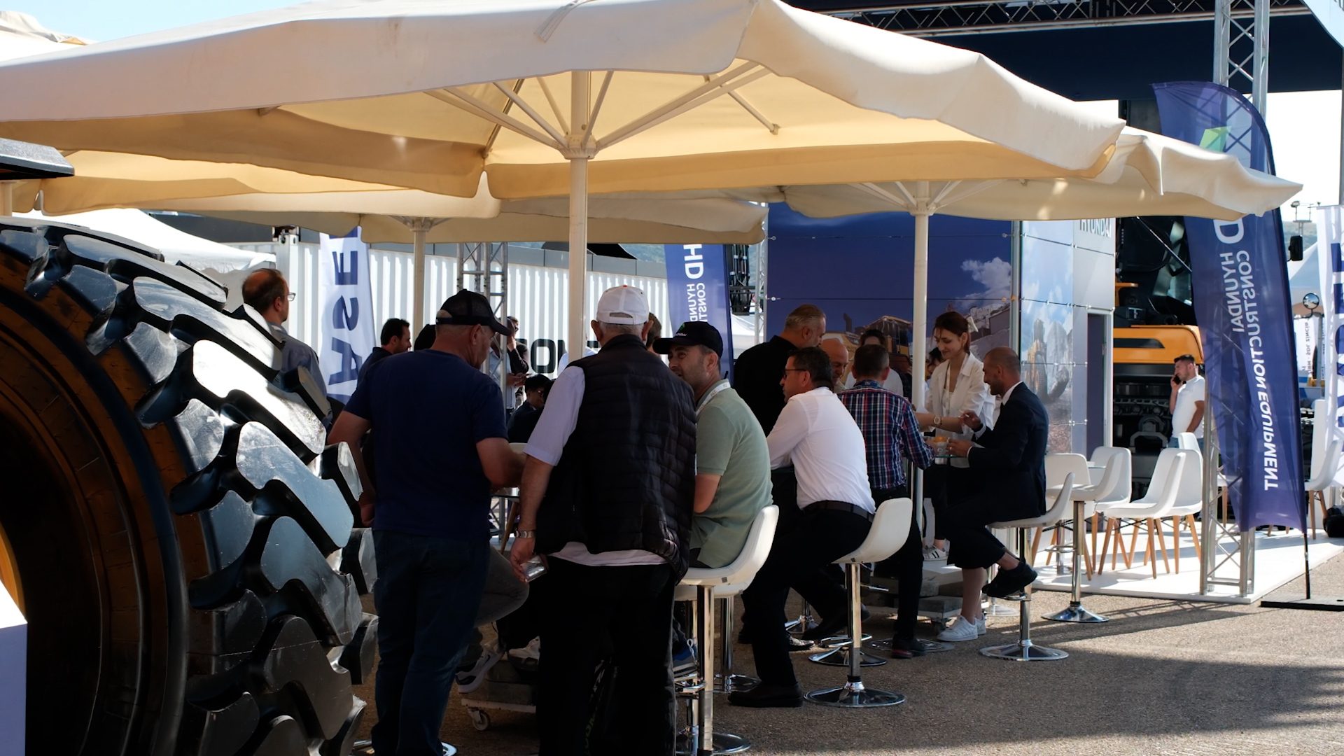 People under umbrellas at a Hyundai Construction Equipment outdoor event, large tire visible.