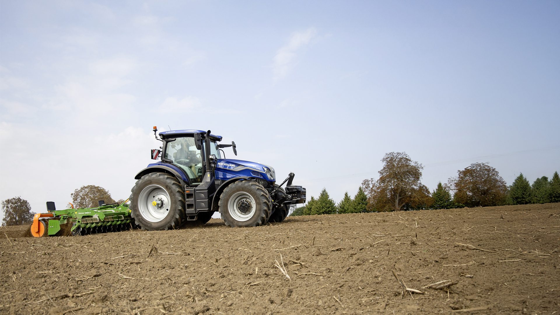 Blue New Holland tractor with a green cultivator tilling a field under a clear sky.