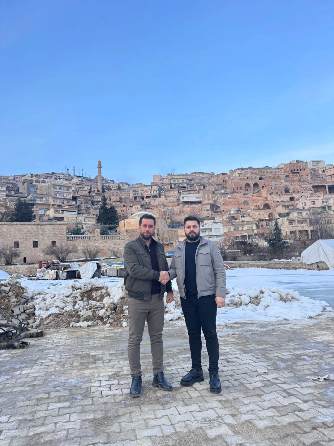 Two men shaking hands in front of a hillside city with stone buildings and snow on the ground.