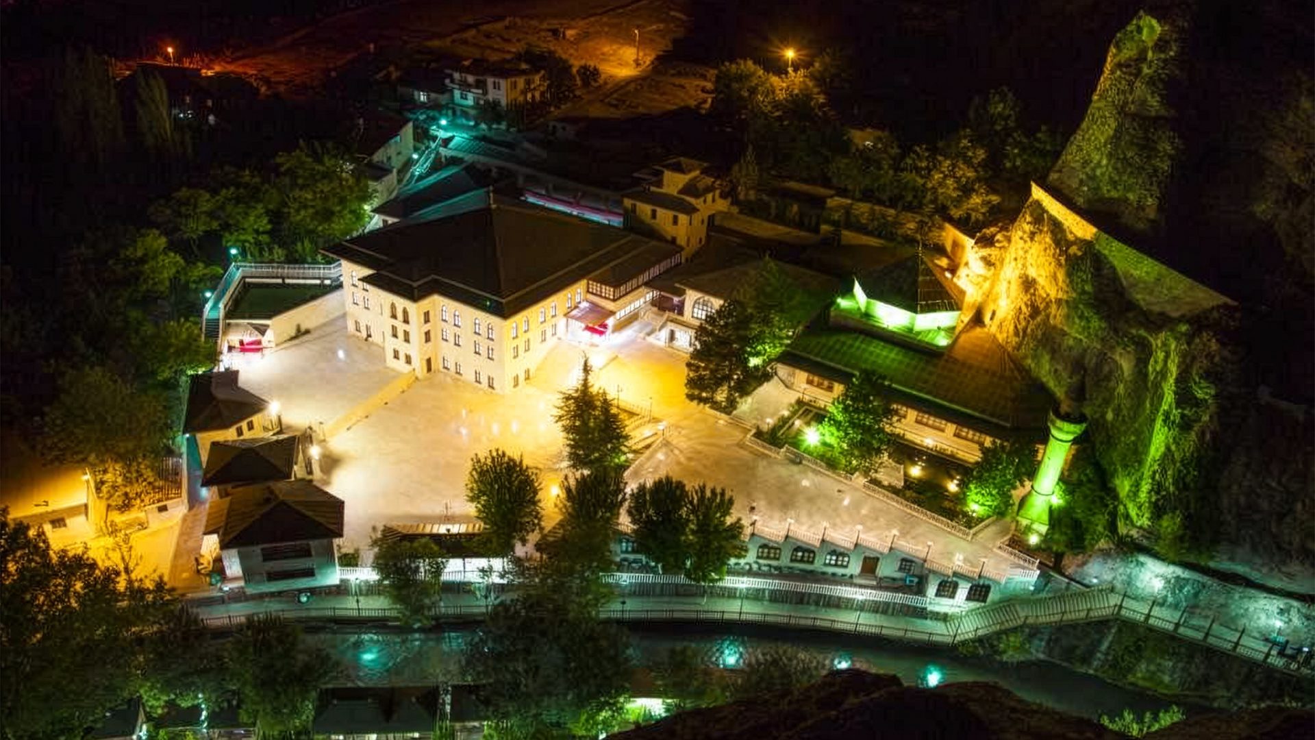 Aerial night view of an illuminated mosque and buildings beside a rocky cliff and river at night.