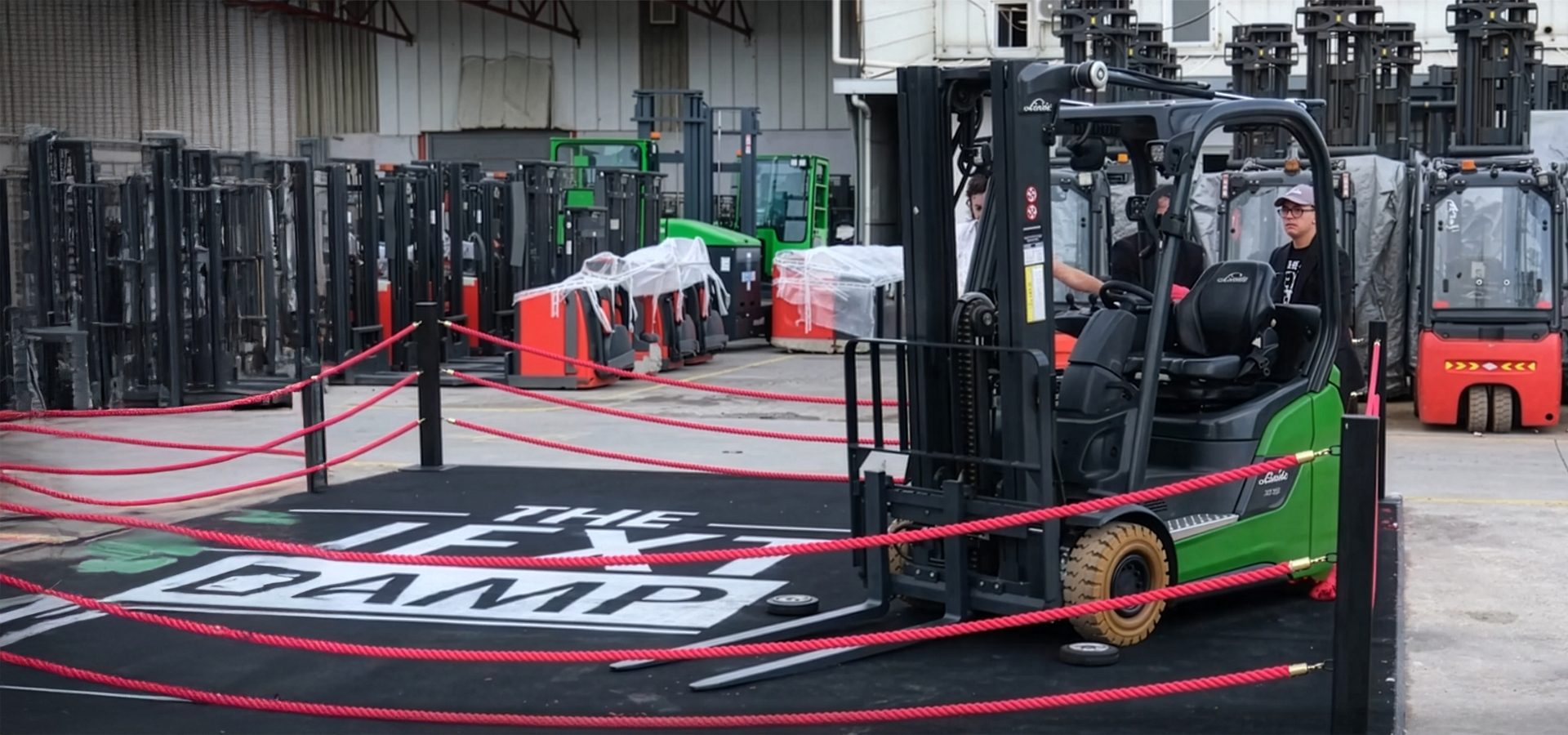 A person sits in a green forklift on a platform resembling a boxing ring, surrounded by other forklifts.