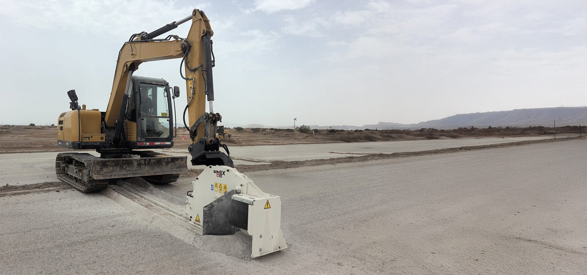 Excavator with SIMEX attachment trenching pavement in an open, barren landscape.
