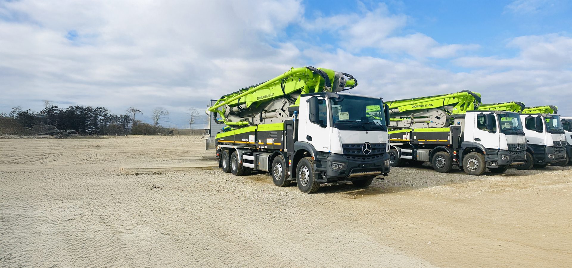 White trucks with green concrete pumps parked on a dirt field under a cloudy sky.