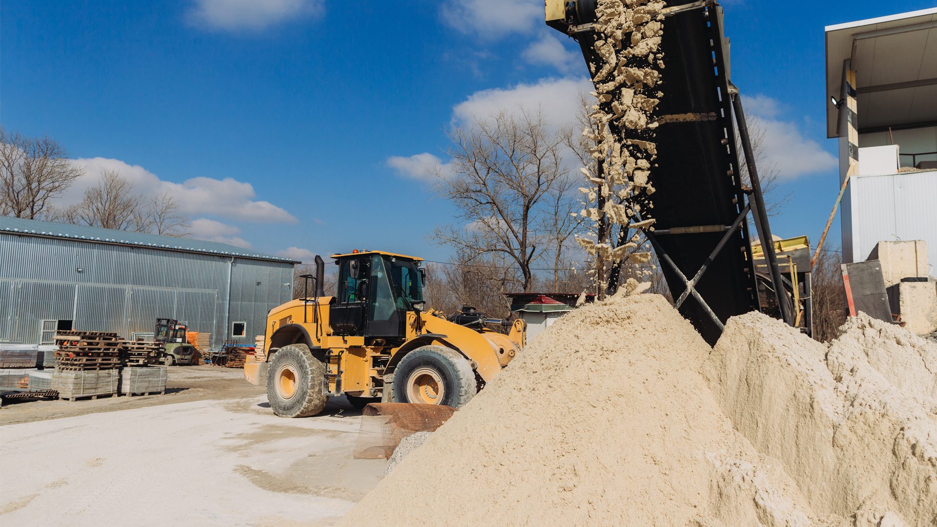 A yellow front-end loader and a conveyor belt dropping material onto a large pile at a processing plant.