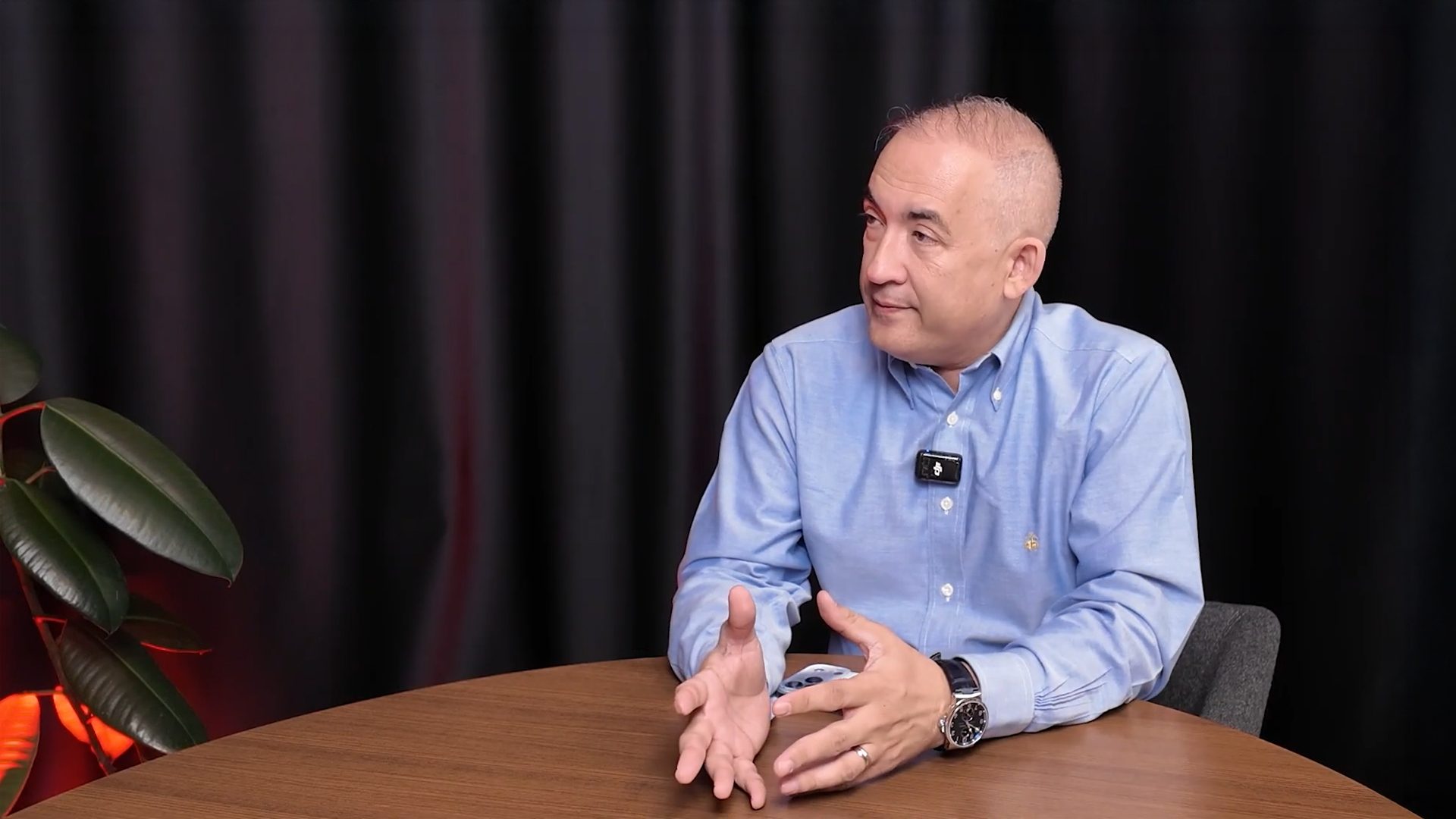 A man in a blue shirt is seated at a table, gesturing with his hands while looking to the right.