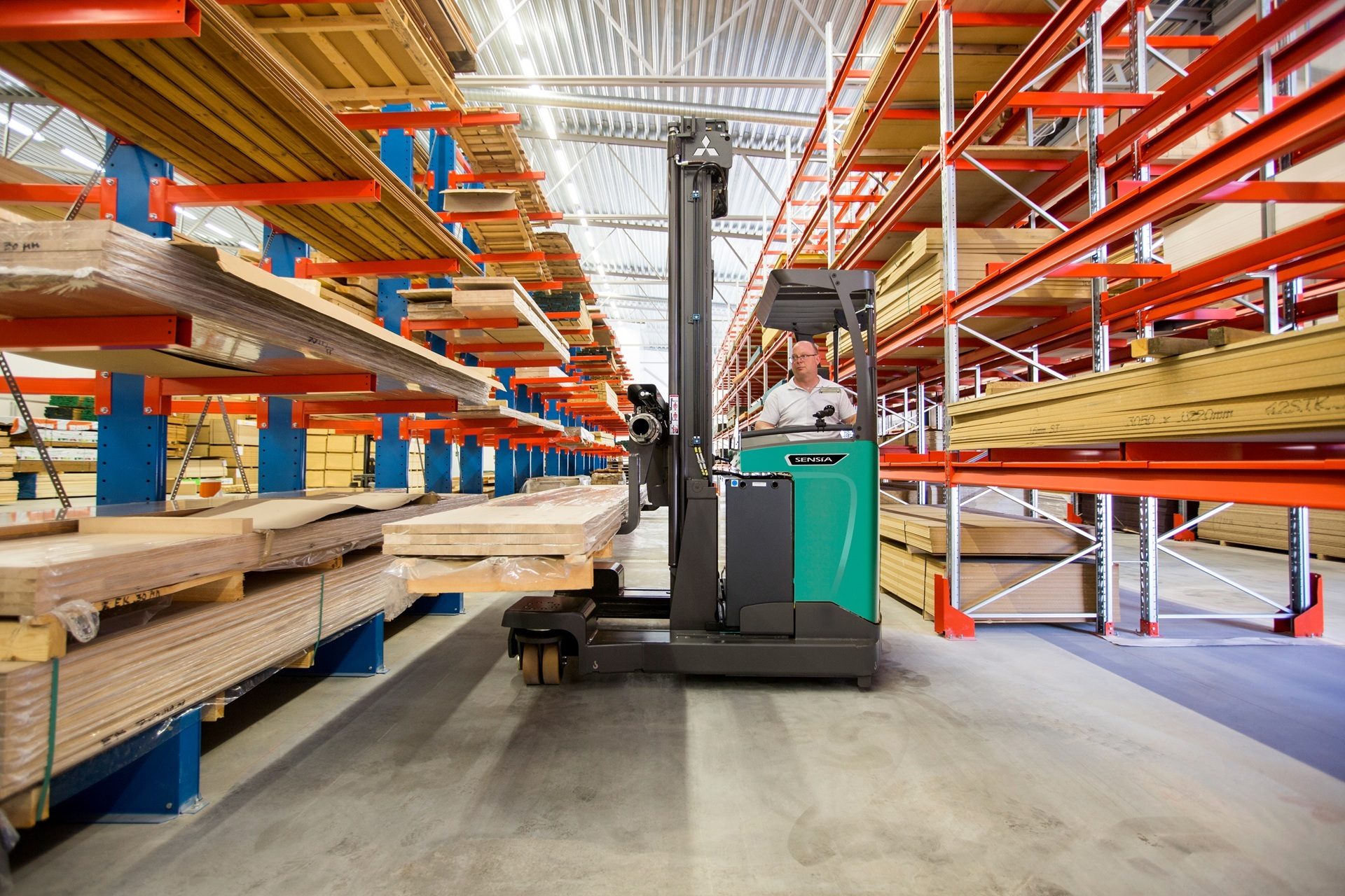 A man operates a green reach truck, moving timber panels in a vast warehouse with high racking.