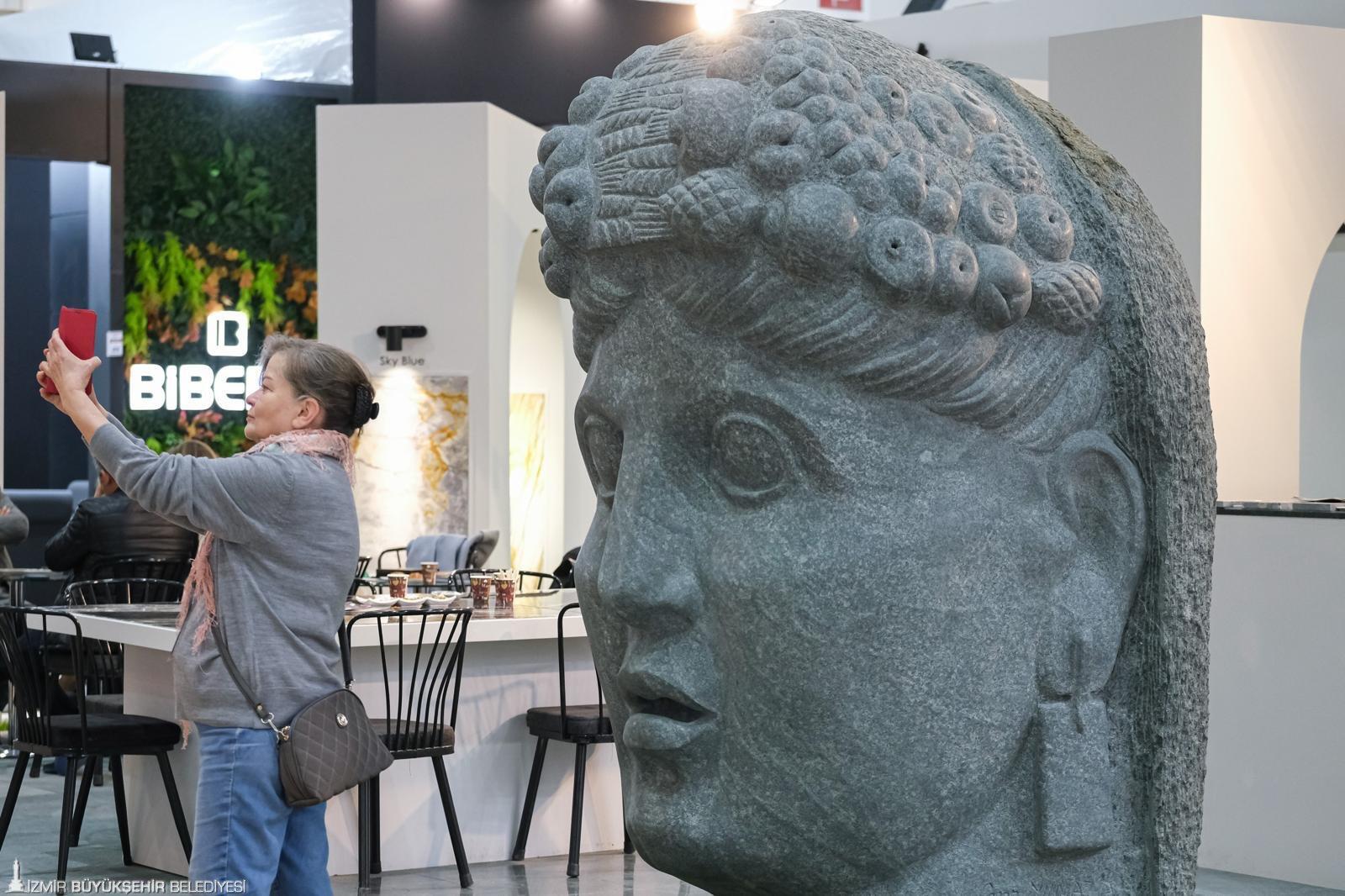 Woman taking a photo of a massive stone head sculpture at an exhibition booth, with green wall in background.