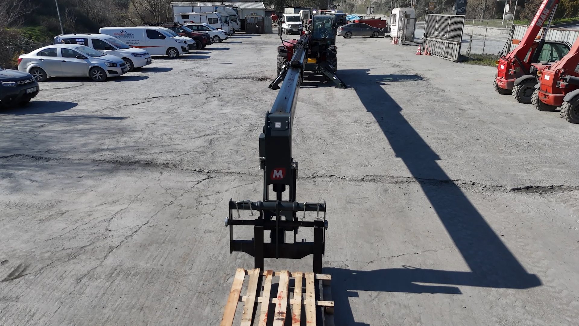 An overhead view of a telehandler with a forklift attachment holding a wooden pallet in an industrial parking lot.
