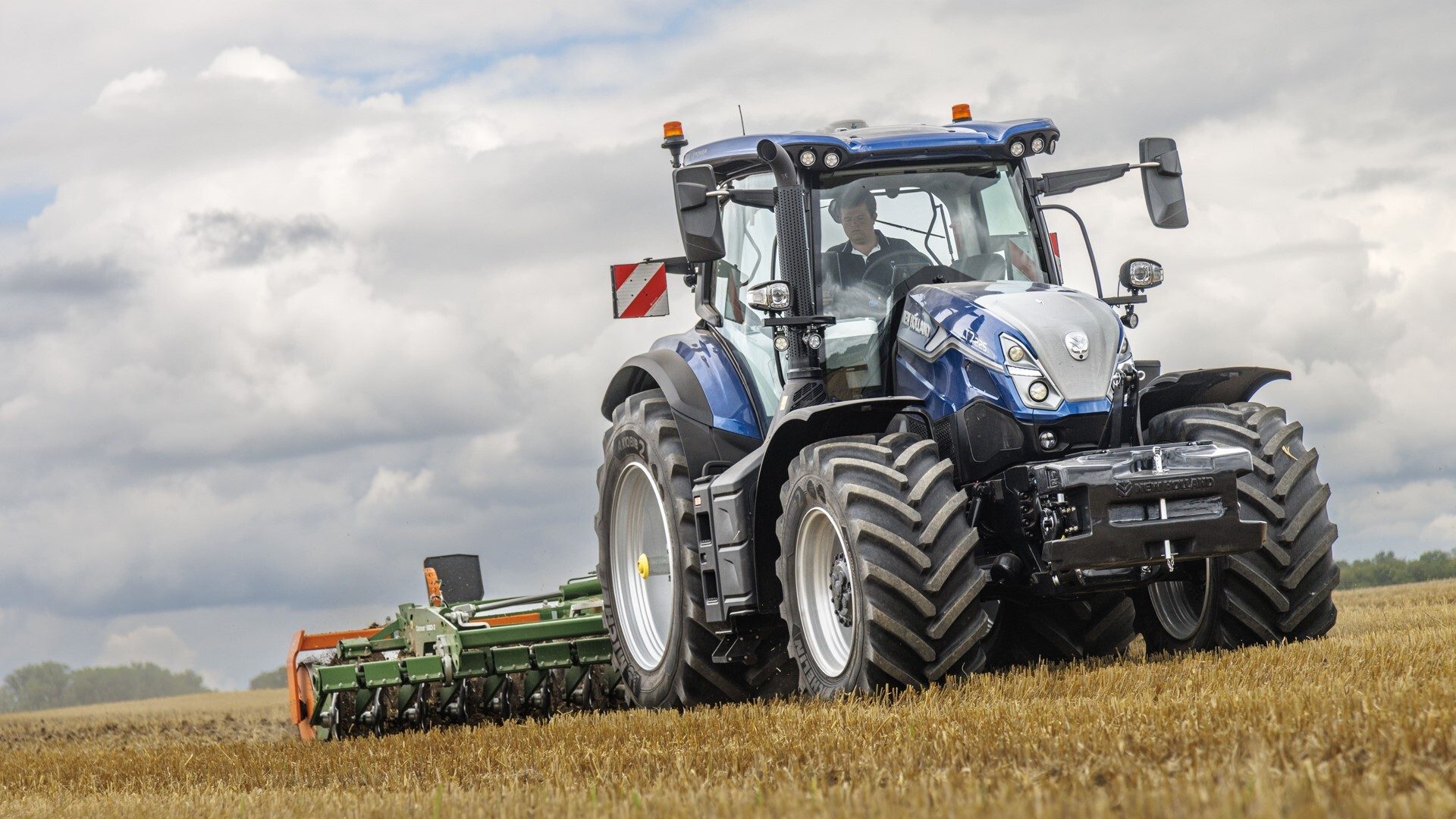 Blue New Holland tractor with driver tilling a golden field under a cloudy sky.
