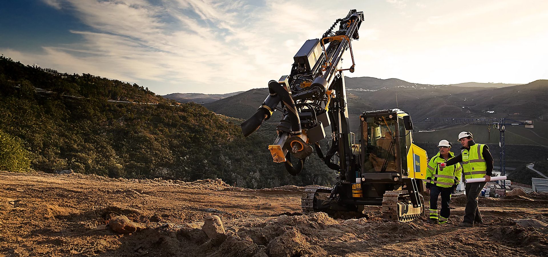 Two workers discuss plans beside a large drilling machine at a mountainous construction site.