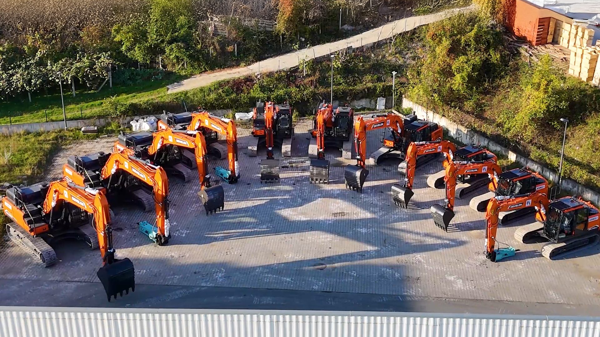 Multiple orange excavators are parked in rows on a paved lot, surrounded by green foliage.