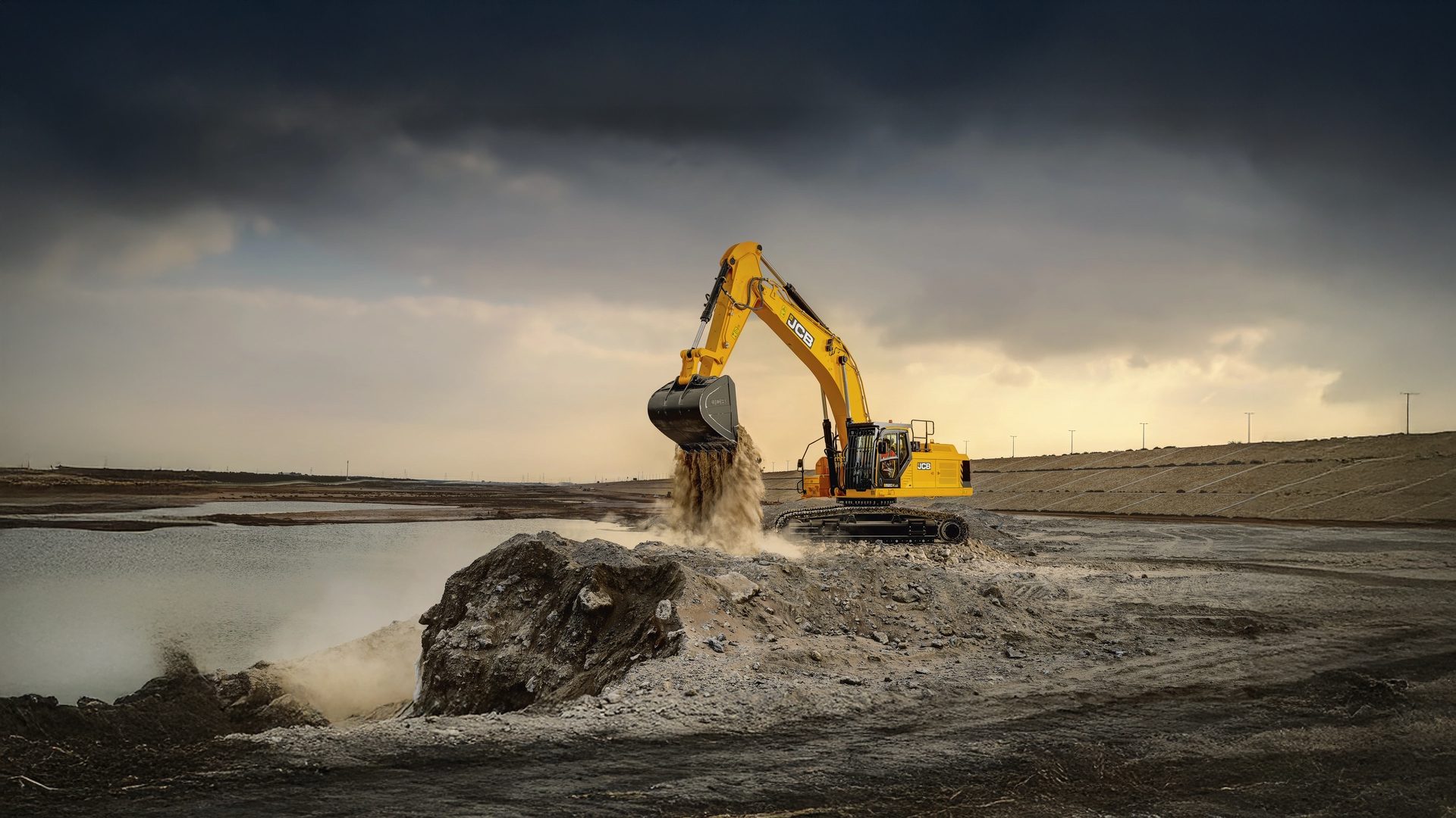 A yellow JCB excavator dumps sand into water under a dark, cloudy sky, creating dust.