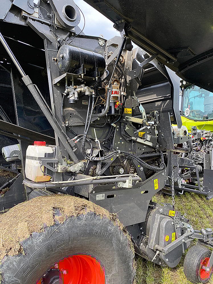 Close-up of a baler's internal mechanisms, including hoses, tanks, gears, and a hay-covered tire.