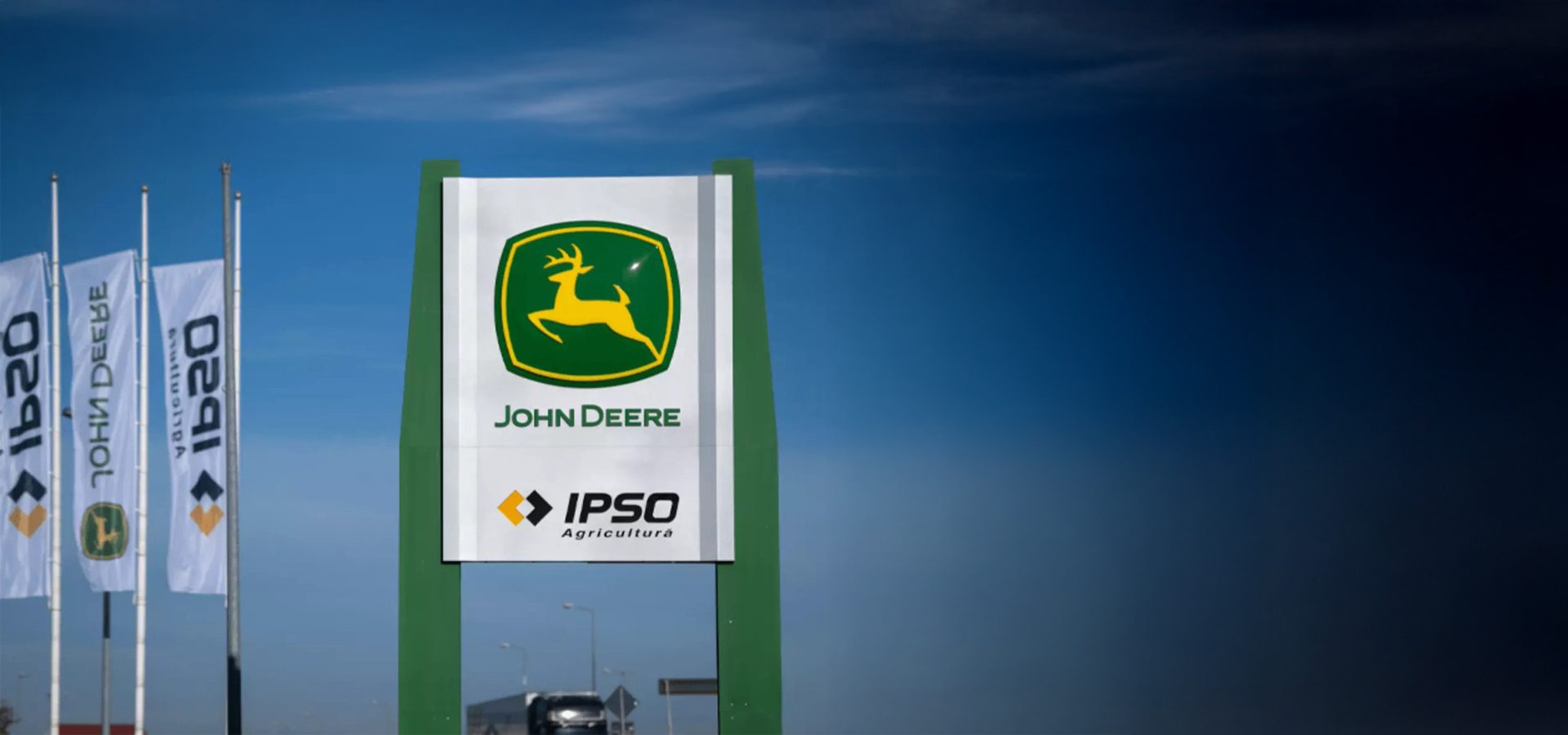 Large John Deere sign and flags under a blue sky, with a road and vehicle.