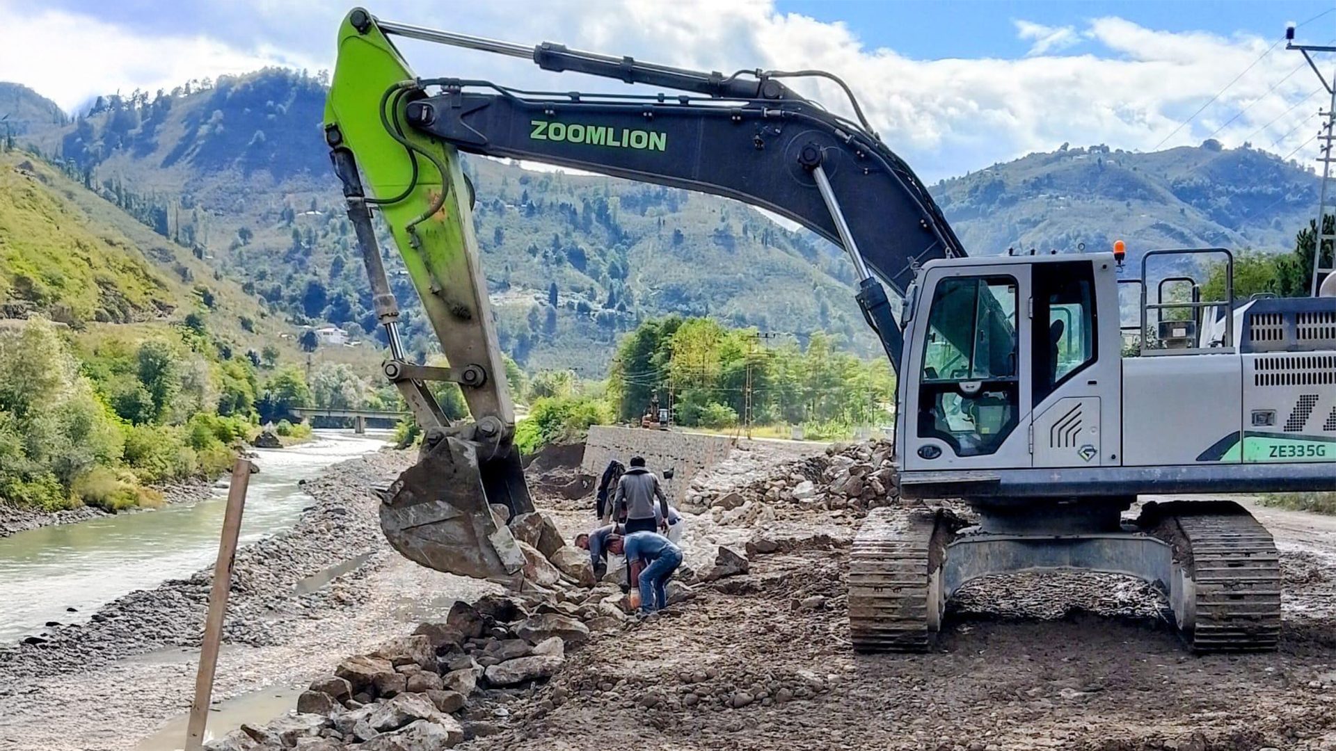 Heavy excavator and workers construct a river bank in a mountainous region.