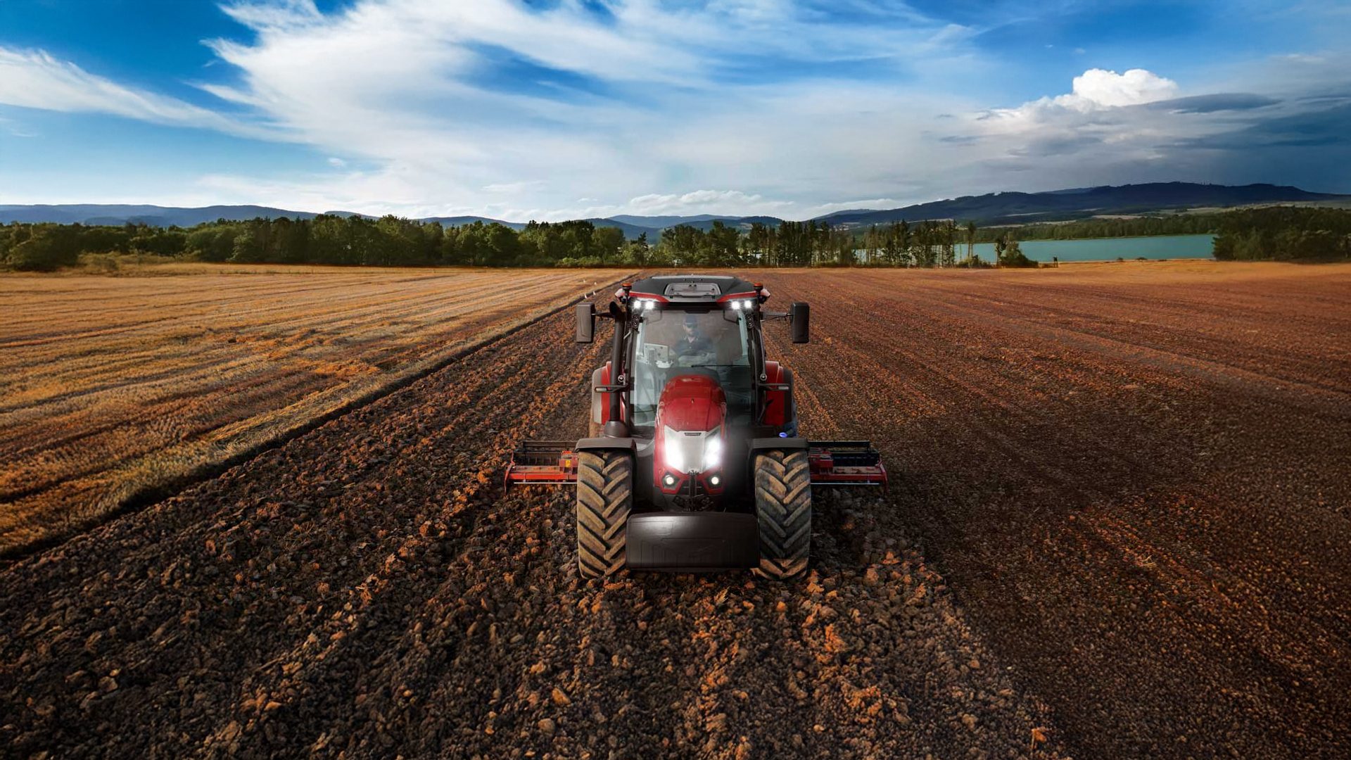Red tractor plowing a field under a blue sky, with a lake and mountains in the background.